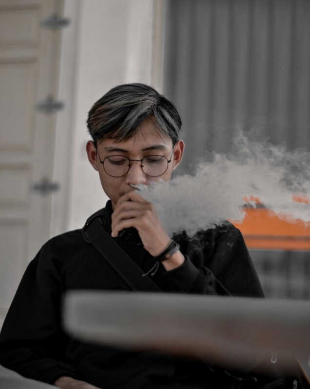 A man wearing glasses is smoking an electronic cigarette, showcasing a relaxed demeanor and modern lifestyle