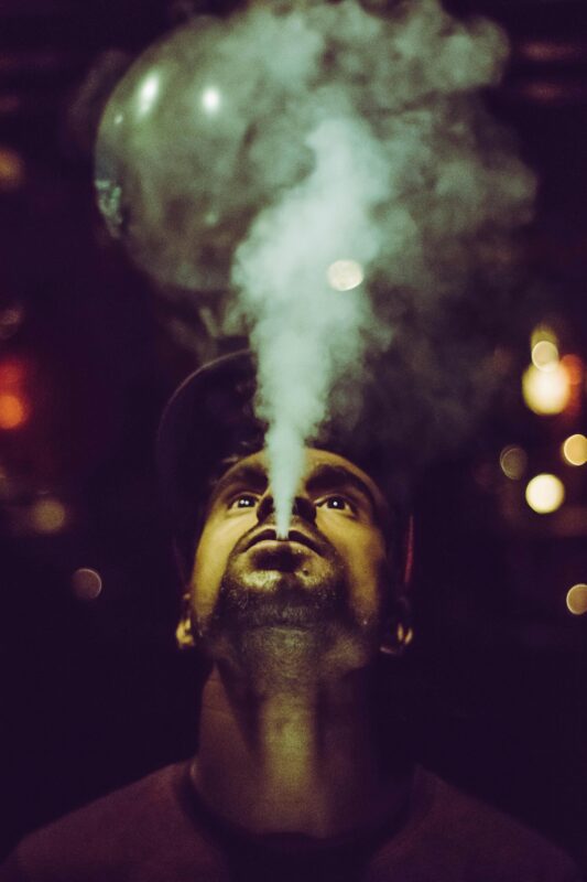 A man exhales smoke, showcasing a relaxed demeanor, with a backdrop hinting at the Backpackboyz brand.